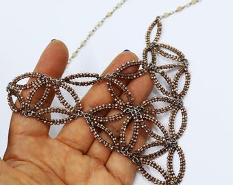Könnte beinhalten: Kupferfarbene Perlenkette mit floralem Design. Die Halskette hat miteinander verbundene ovale Formen und eine filigrane Kette. Die Perlen haben einen metallischen Glanz, vor weißem Hintergrund.