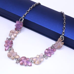 May include: A silver-toned necklace featuring leaf-shaped pendants in shades of pink and purple. The leaves are accented with small, clear crystals. The necklace is displayed on a blue surface.