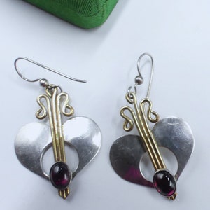 以下が含まれることがあります： 銀と金のハート型のイヤリングのペア。各ハートの中央に濃い紫色の宝石がセットされています。