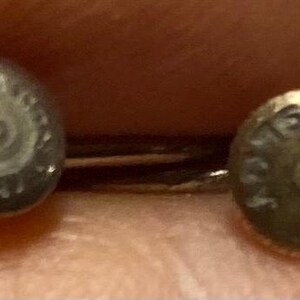 K&ouml;nnte beinhalten: Nahaufnahme von zwei kleinen, silberfarbenen Ohrringen. Jeder Ohrring hat ein kreisf&ouml;rmiges Design mit ge&auml;tzten Details. Die Ohrringe sind vor einem verschwommenen Hintergrund platziert, was darauf hindeutet, dass sie gehalten werden.