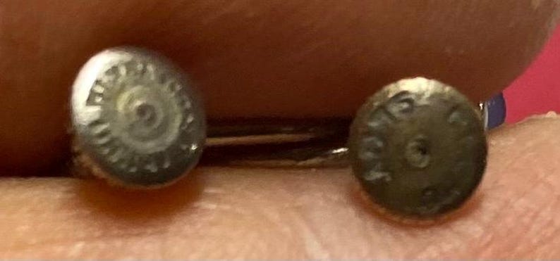 K&ouml;nnte beinhalten: Nahaufnahme von zwei kleinen, silberfarbenen Ohrringen. Jeder Ohrring hat ein kreisf&ouml;rmiges Design mit ge&auml;tzten Details. Die Ohrringe sind vor einem verschwommenen Hintergrund platziert, was darauf hindeutet, dass sie gehalten werden.