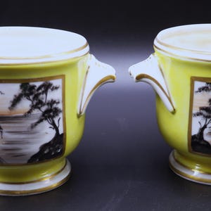 May include: A pair of yellow porcelain planters with white rims and handles. Each planter has a rectangular landscape scene with trees, water, and a building. The planters have gold trim and a pedestal base.