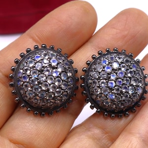 May include: A pair of round stud earrings with a dark metal setting. Each earring is encrusted with small, clear and blue gemstones. The earrings have a beaded edge and are displayed on a light background.