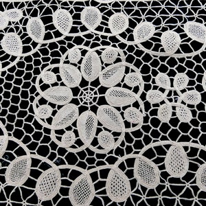 May include: White crocheted lace fabric with a delicate floral pattern. The lace has a fine, intricate design with small, oval-shaped motifs.