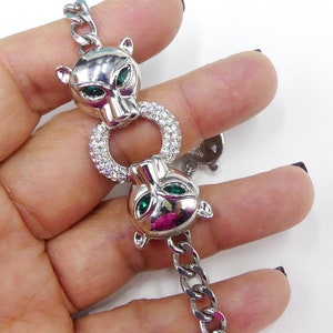 Könnte beinhalten: Silberfarbenes Armband mit zwei Pantherkopf-Anhängern mit grünen Augen, verbunden durch einen Ring aus klaren Kristallen. Das Armband hat ein Kettendesign. Es ist ein Schmuckstück.
