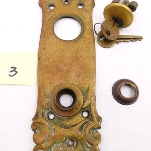 May include: Antique brass door plate with ornate floral design and a keyhole. The plate has a worn, aged finish and is accompanied by a Yale key and a separate brass escutcheon.