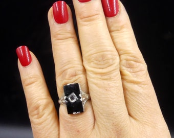 May include: A silver ring with a black rectangular stone set in a filigree design. The stone has a small diamond-shaped clear stone in the center.