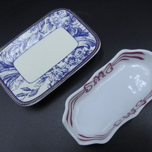 May include: Two ceramic serving dishes. One is rectangular with a white centre and a blue floral design border with gold trim. The other is oval-shaped with a white interior and a burgundy decorative border.