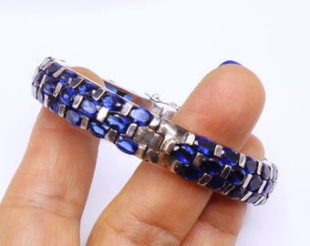 May include: A silver bracelet with three rows of oval, deep blue gemstones. The bracelet is held in a hand against a white background, showcasing the jewelry's design and color.