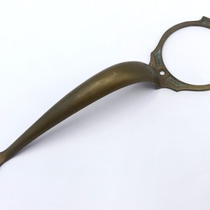 May include: A brass door handle with an ornate design. The handle has a long, curved arm and a circular ring at the end. The handle is a vintage style.