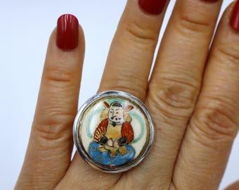 May include: A silver ring with a round, colorful enamel inlay depicting a person in traditional clothing. The enamel is blue, red, yellow, and white.
