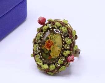 May include: Oval-shaped ring with a floral design. The ring features a central image of roses, surrounded by green ribbon, rhinestones, and small red flower accents. The ring has a bronze-colored band.
