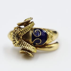 May include: A gold ring featuring a blue enamel design and a detailed ram's head. The ram's horns are curled and the enamel is a deep blue with gold accents.