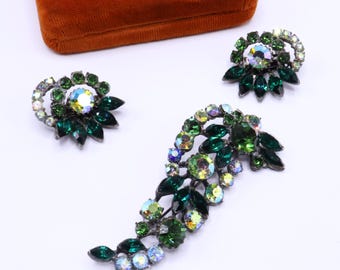 May include: A vintage jewelry set featuring a brooch and matching earrings. The set showcases a floral design with emerald green and iridescent crystals. The brooch has a curved shape, and the earrings are circular. The jewelry is set against a white background.