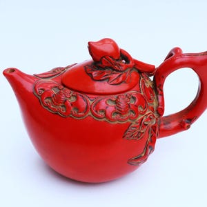 May include: A red ceramic teapot with intricate carvings and a detailed lid. The teapot has a rounded body and a curved handle.