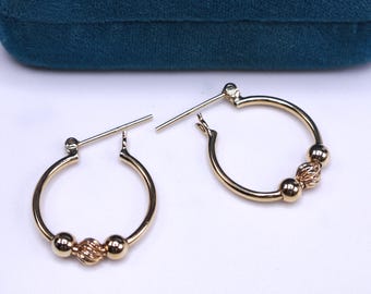 May include: A pair of gold hoop earrings. Each earring features a small, textured bead between two smooth, round beads. The earrings are displayed on a white surface with a dark teal velvet box in the background.