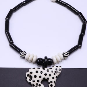 May include: A black and white beaded necklace with a spotted cat pendant. The necklace features black and white cylindrical beads, white round beads, and a black cat pendant with black spots. The necklace is on a white and black background.