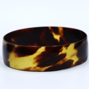 May include: A wide, oval bangle bracelet with a tortoiseshell pattern. The bracelet has a mix of dark brown and golden-yellow hues, creating a marbled effect. The smooth, polished surface reflects light, highlighting the colour variations.