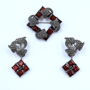 May include: A set of silver earrings and a brooch with a square design. The jewelry features a dark gray metal with red gemstones.