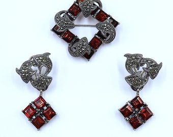May include: A set of silver earrings and a brooch with a square design. The jewelry features a dark gray metal with red gemstones.