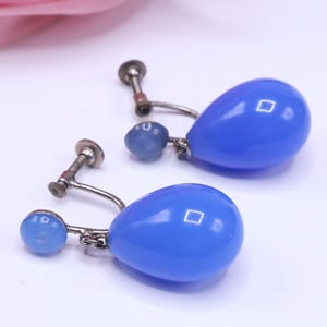 K&ouml;nnte beinhalten: Ein Paar Vintage-Ohrringe in Tropfenform in Blau. Jeder Ohrring hat eine gro&szlig;e, gl&auml;nzende, blaue, tropfenf&ouml;rmige Perle, die an einem silberfarbenen Clip-On-Verschluss befestigt ist. Eine kleinere blaue Perle ist ebenfalls angebracht.