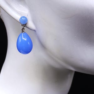 K&ouml;nnte beinhalten: Ein Paar hellblaue Ohrringe. Jeder Ohrring hat einen kleinen, runden Stecker und einen gr&ouml;&szlig;eren, tropfenf&ouml;rmigen Anh&auml;nger. Die Ohrringe sind in einem leuchtenden Blauton gehalten und scheinen gl&auml;nzend zu sein. Sie werden auf einer wei&szlig;en Mannequin pr&auml;sentiert.