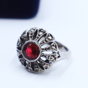 Puede incluir: Un anillo de plata con una piedra preciosa roja rodeada de un delicado diseño de filigrana.