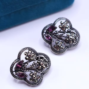May include: Two silver-toned floral earrings with marcasite accents and oval-cut purple gemstones. The earrings have a clover-like shape with detailed filigree work. The background is white with a teal velvet jewelry box.