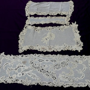 May include: A collection of antique white lace textiles, including a long runner and several smaller pieces. The lace features intricate floral and scrollwork patterns, with scalloped edges. The textiles are arranged on a dark purple background.