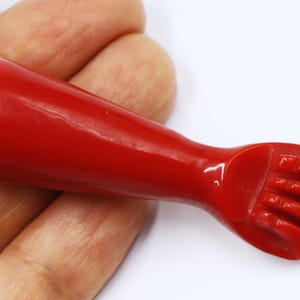 Puede incluir: Un brazo de plástico rojo con una mano, la mano está en forma de puño con el pulgar extendido. El brazo mide aproximadamente 10 cm de largo y tiene un pasador de metal en la parte superior.