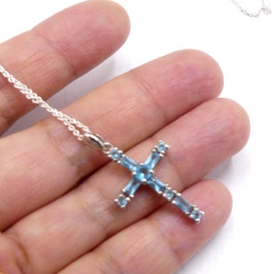 May include: A silver chain necklace with a blue topaz cross pendant. The cross is set with small, rectangular-shaped stones.