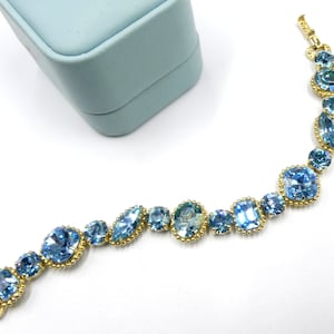 May include: A gold bracelet with a chain and blue crystal stones. The bracelet is on a light blue background.