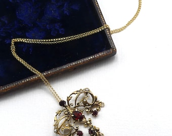 May include: A gold necklace with a pendant featuring a delicate, ornate design with red and white gemstones. The pendant is shaped like a teardrop with a blue gemstone at the bottom.