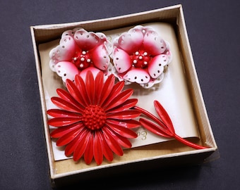 May include: A set of vintage floral brooches and earrings in a box. The earrings are white with red accents, and the brooch is a vibrant red daisy. The box is beige with a dark background.