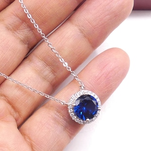 May include: A silver chain necklace with a pendant featuring a large, round, dark blue gemstone surrounded by a halo of smaller, clear gemstones.