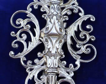 May include: A silver filigree brooch with a detailed floral design. The brooch is made of intricate swirls and loops, creating a delicate and elegant piece.