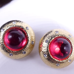 以下が含まれることがあります： 赤いガラスのカボションとクリアなラインストーンが付いた、ゴールドトーンのクリップ式イヤリングのペアです。