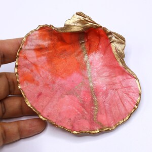 Op de afbeelding: Een decoratief schelpvormig schaaltje met een goud geschilderde rand en accenten. De binnenkant heeft een mix van roze en oranje tinten, wat een levendige en artistieke uitstraling creëert. Het schaaltje wordt door een hand tegen een witte achtergrond gehouden.