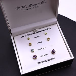 May include: An open jewelry box displaying a set of stud earrings. The earrings are gold-colored and feature various gemstones, including peridot, citrine, blue topaz, and amethyst. The box is labeled "R.H. Macy & Co. FINE JEWELRY".