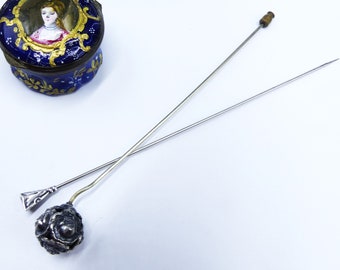 May include: Two antique hat pins with silver and brass accents. One pin features a decorative silver floral design, while the other has a geometric silver top. A small, blue, decorative trinket box with a portrait on the lid is also visible.