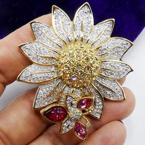 May include: A gold-tone brooch featuring a large sunflower with a yellow centre and a smaller butterfly with pink and red accents. The sunflower and butterfly are both covered in clear rhinestones.