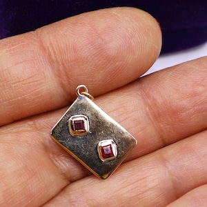 May include: A gold-toned pendant with a diamond shape, featuring two square, red gemstones. The pendant is held in a hand, showcasing its size and design. The jewelry piece has a small loop at the top for a chain.