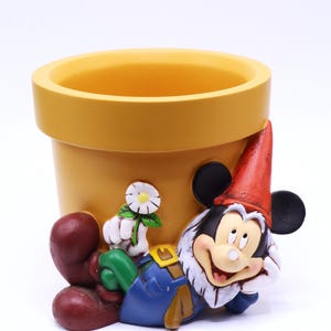 May include: A yellow ceramic flower pot with a smiling Mickey Mouse gnome figure. The gnome wears a red hat, blue overalls, and a white beard. A white daisy is attached to the pot.