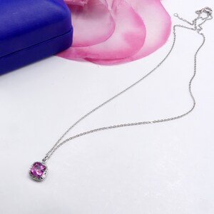 May include: A silver chain necklace with a square-shaped pendant featuring a pink gemstone surrounded by smaller clear gemstones.