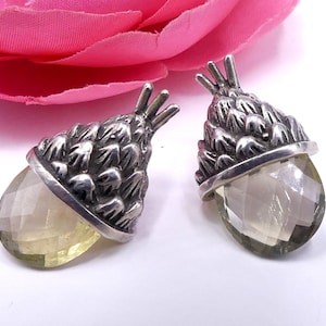 May include: A pair of silver earrings with a clear, faceted gemstone in the shape of an acorn. The acorn top is detailed with a textured, pinecone-like design.