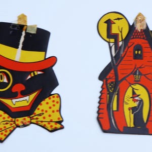 May include: Two vintage Halloween decorations. One is a black cat with a top hat, monocle, and bow tie. The other is a red house with a witch in the doorway and a full moon.