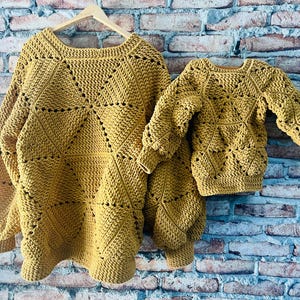 May include: Mustard yellow crochet sweaters, one adult size and one child size, hanging on a wooden hanger. The sweaters have a geometric design with openwork details. The background is a brick wall.