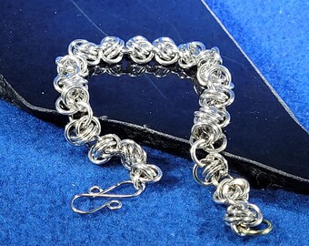 May include: A silver chain bracelet with a unique knot design. The bracelet is lying on a black surface.