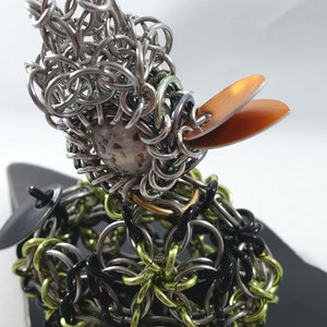 Può includere: Una scultura in metallo argentata, nera e verde di un'anatra con un becco arancione. L'anatra è realizzata in maglia metallica e presenta un design dettagliato.
