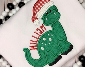 May include: A green dinosaur wearing a red and white gingham Santa hat with white trim. The dinosaur has a white name on its back that says "William".